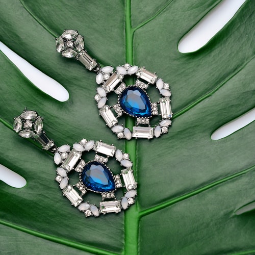 Statement earrings with sapphire-blue crystal centers and white rhinestones on a monstera leaf